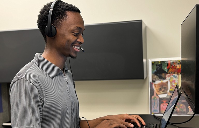 Computer engineering co-op student Ivan Senga provides support for faculty and staff at the CommUNITY Support Centre Young smiling man, seen from the side, working on a desktop computer.